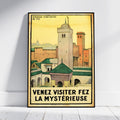 A framed vintage travel poster of Fez, Morocco, leaning against a white wall on a light wooden floor. The poster features a yellow sky and architectural details of the city with the French text 'VENEZ VISITER FEZ LA MYSTÉRIEUSE' at the bottom.