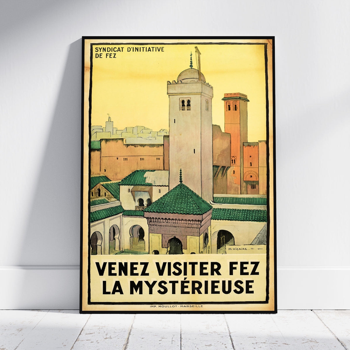 A framed vintage travel poster of Fez, Morocco, leaning against a white wall on a light wooden floor. The poster features a yellow sky and architectural details of the city with the French text 'VENEZ VISITER FEZ LA MYSTÉRIEUSE' at the bottom.