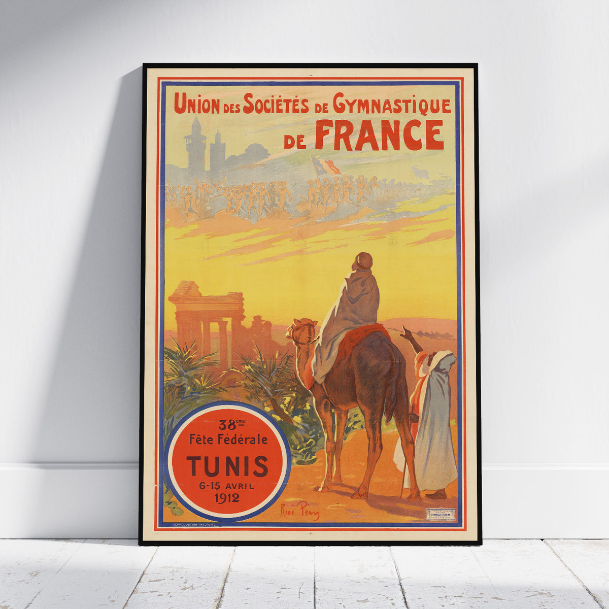 A framed vintage-style poster leaning against a white wall on a light wood floor. The poster features illustrated figures, camels, and architectural elements with the text 'Union des Sociéts de Gymnastique de France' and 'Tunis' in a red circular badge.