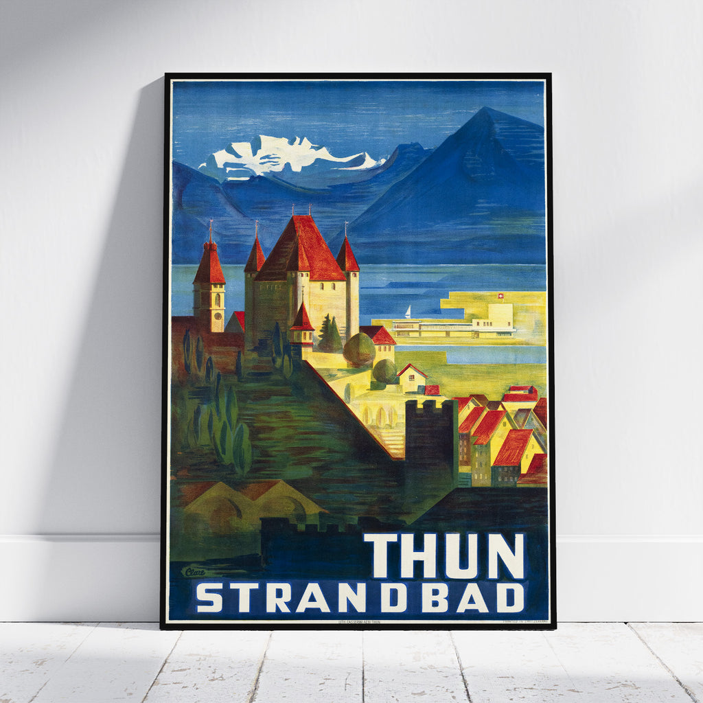 A framed vintage-style travel poster leaning against a white wall on a light wood floor. The poster depicts a coastal town with red-roofed buildings, blue mountains, and the text 'THUN STRANDBAD' at the bottom.