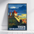 A framed vintage-style travel poster leaning against a white wall on a light wood floor. The poster depicts a coastal town with red-roofed buildings, blue mountains, and the text 'THUN STRANDBAD' at the bottom.