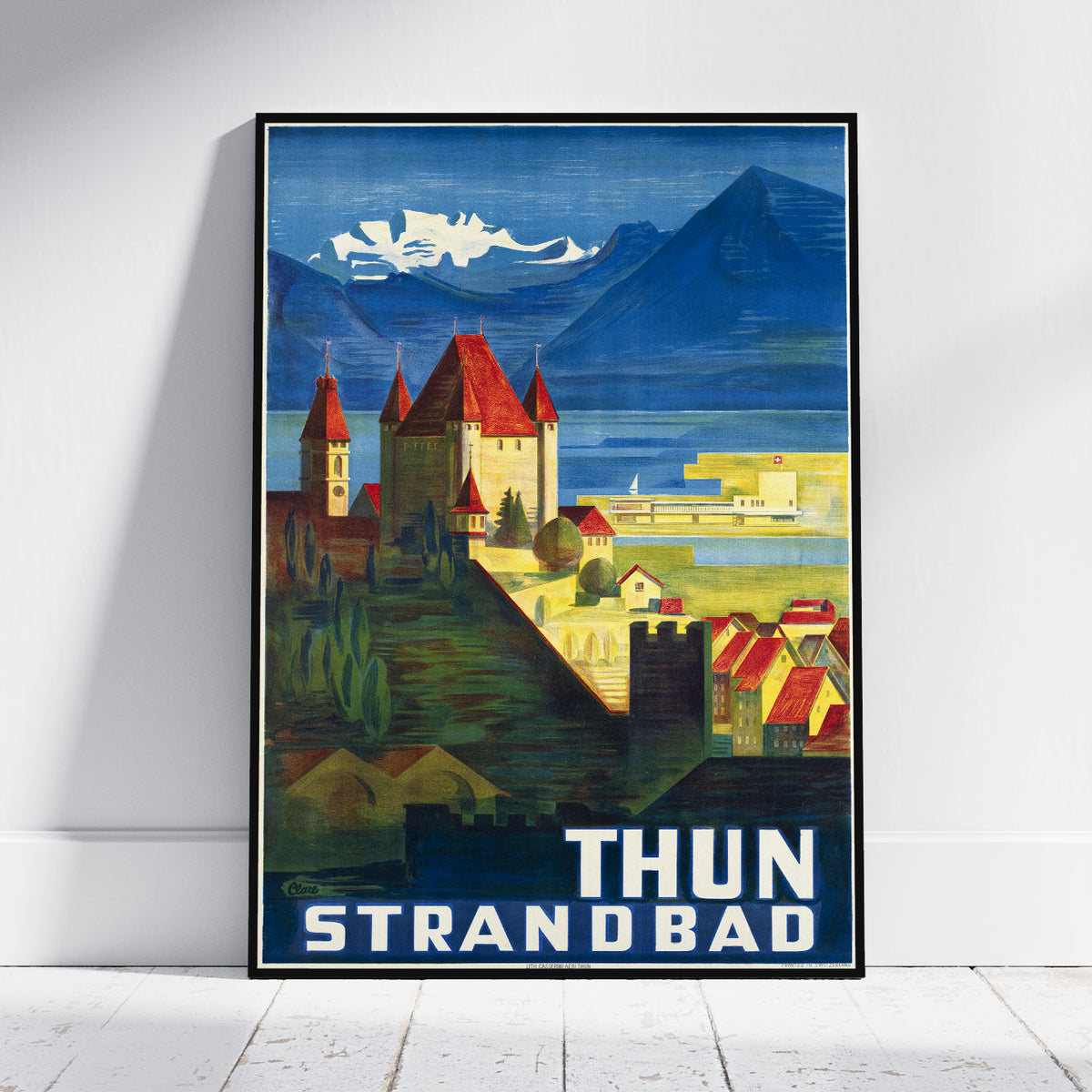 A framed vintage-style travel poster leaning against a white wall on a light wood floor. The poster depicts a coastal town with red-roofed buildings, blue mountains, and the text 'THUN STRANDBAD' at the bottom.