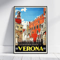A framed vintage-style travel poster of Verona, Italy, leaning against a white wall on a light wood floor. The poster features a colorful illustration of the city's architecture, including a prominent red-orange building, a bell tower, and a blue sky, with 'VERONA' printed in bold yellow letters at the bottom.