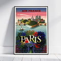 A framed vintage-style Paris travel poster leaning against a white wall on a light floor. The poster features the text 'AIR FRANCE' at the top and 'PARIS' in large letters over a colorful illustration of the city skyline, water, and foliage.