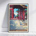 A framed art print featuring a Japanese-style scene with red temple structures, stone steps, and a figure in blue, leaning against a white wall on a light floor.