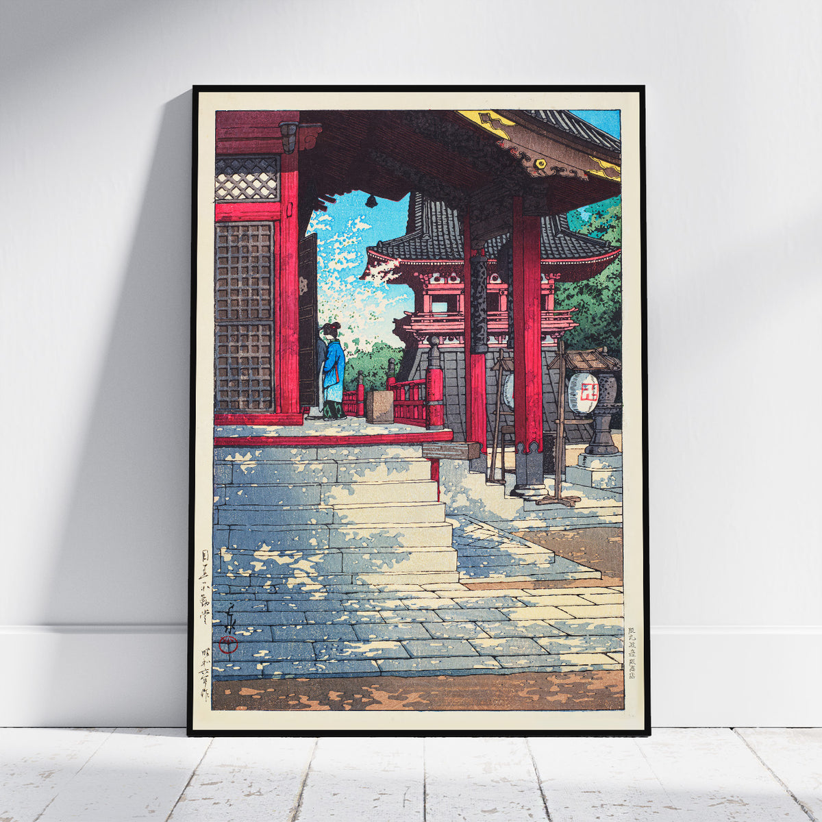 A framed art print featuring a Japanese-style scene with red temple structures, stone steps, and a figure in blue, leaning against a white wall on a light floor.