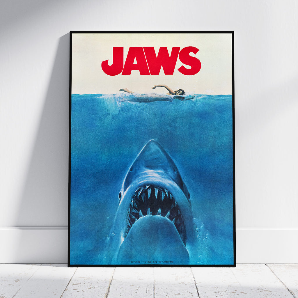 A framed Jaws movie poster leaning against a white wall on a light wood floor. The poster features the title 'JAWS' in bold red letters above a blue underwater scene with a shark rising toward the surface and a swimmer at the waterline.