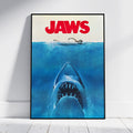 A framed Jaws movie poster leaning against a white wall on a light wood floor. The poster features the title 'JAWS' in bold red letters above a blue underwater scene with a shark rising toward the surface and a swimmer at the waterline.