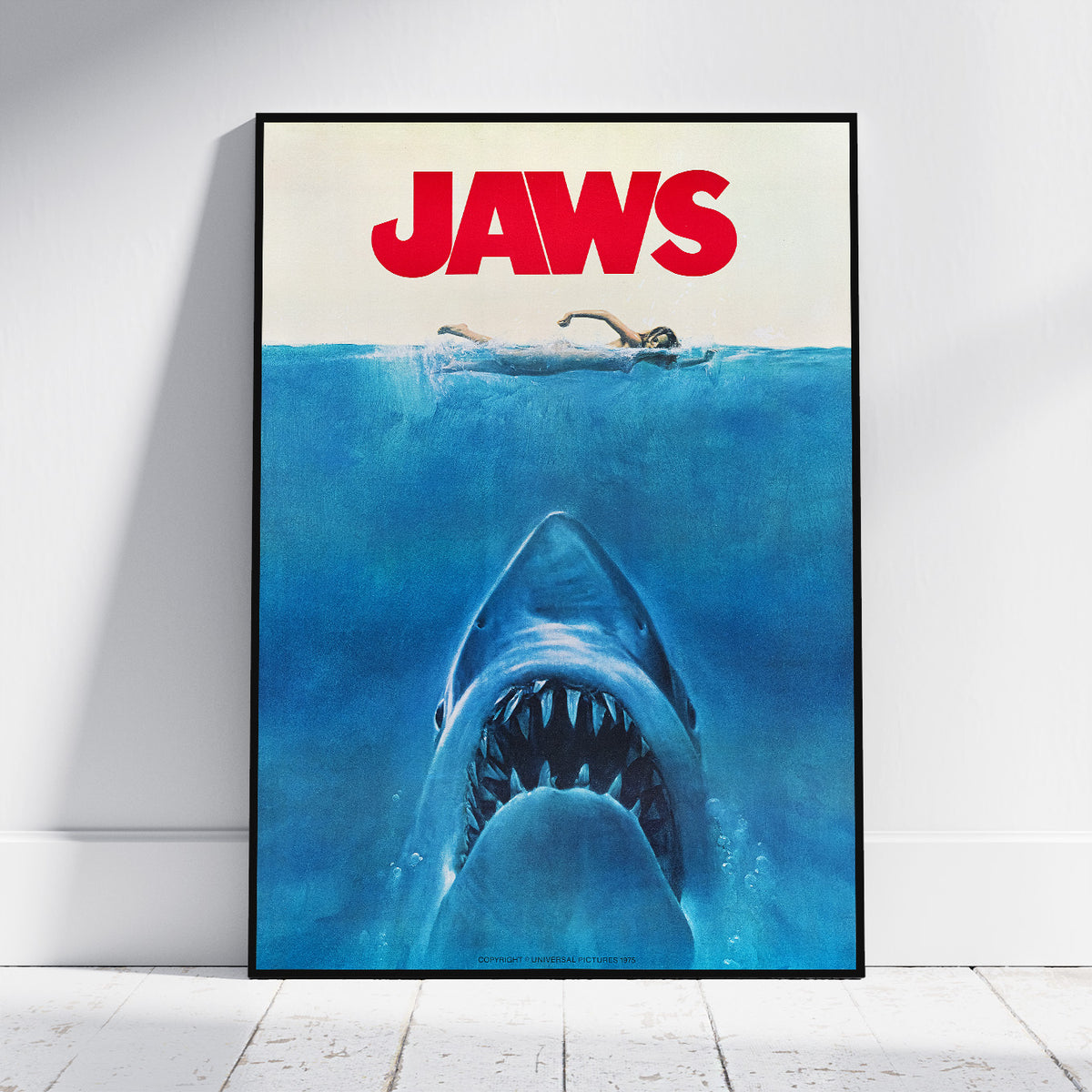 A framed Jaws movie poster leaning against a white wall on a light wood floor. The poster features the title 'JAWS' in bold red letters above a blue underwater scene with a shark rising toward the surface and a swimmer at the waterline.