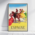A framed vintage-style travel poster leaning against a white wall on a light wood floor. The poster depicts Spanish dancers and riders with large 'ESPAGNE' text at the bottom.