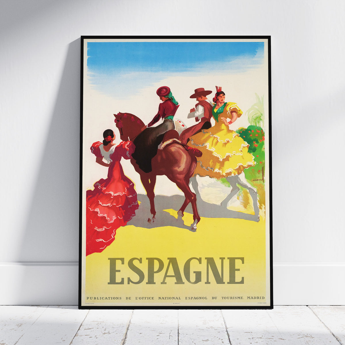 A framed vintage-style travel poster leaning against a white wall on a light wood floor. The poster depicts Spanish dancers and riders with large 'ESPAGNE' text at the bottom.