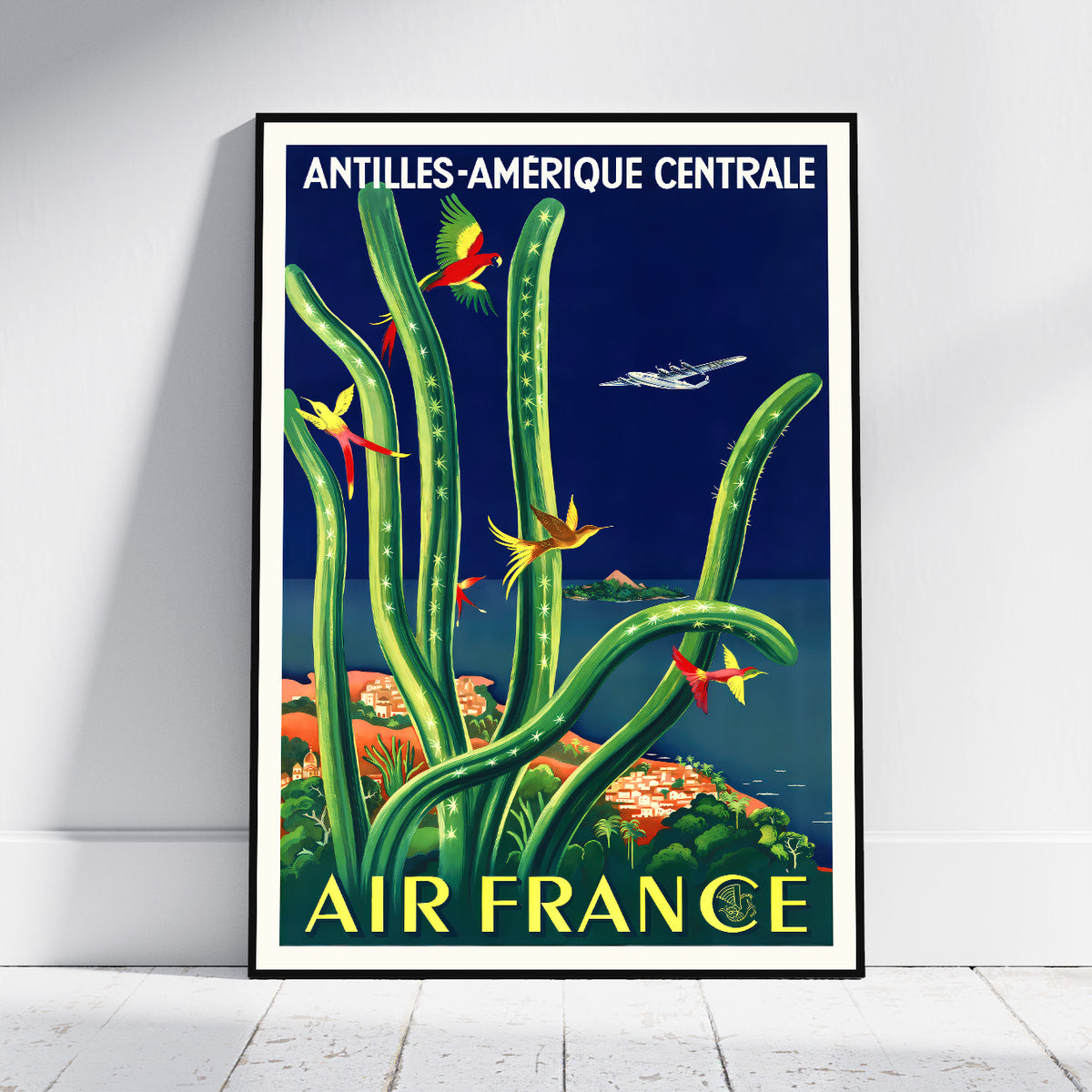A framed vintage-style travel poster leaning against a white wall on a light wood floor. The poster features tall green cacti, colorful birds, an airplane, and the text 'ANTILLES-AMERIQUE CENTRALE' at the top and 'AIR FRANCE' at the bottom.