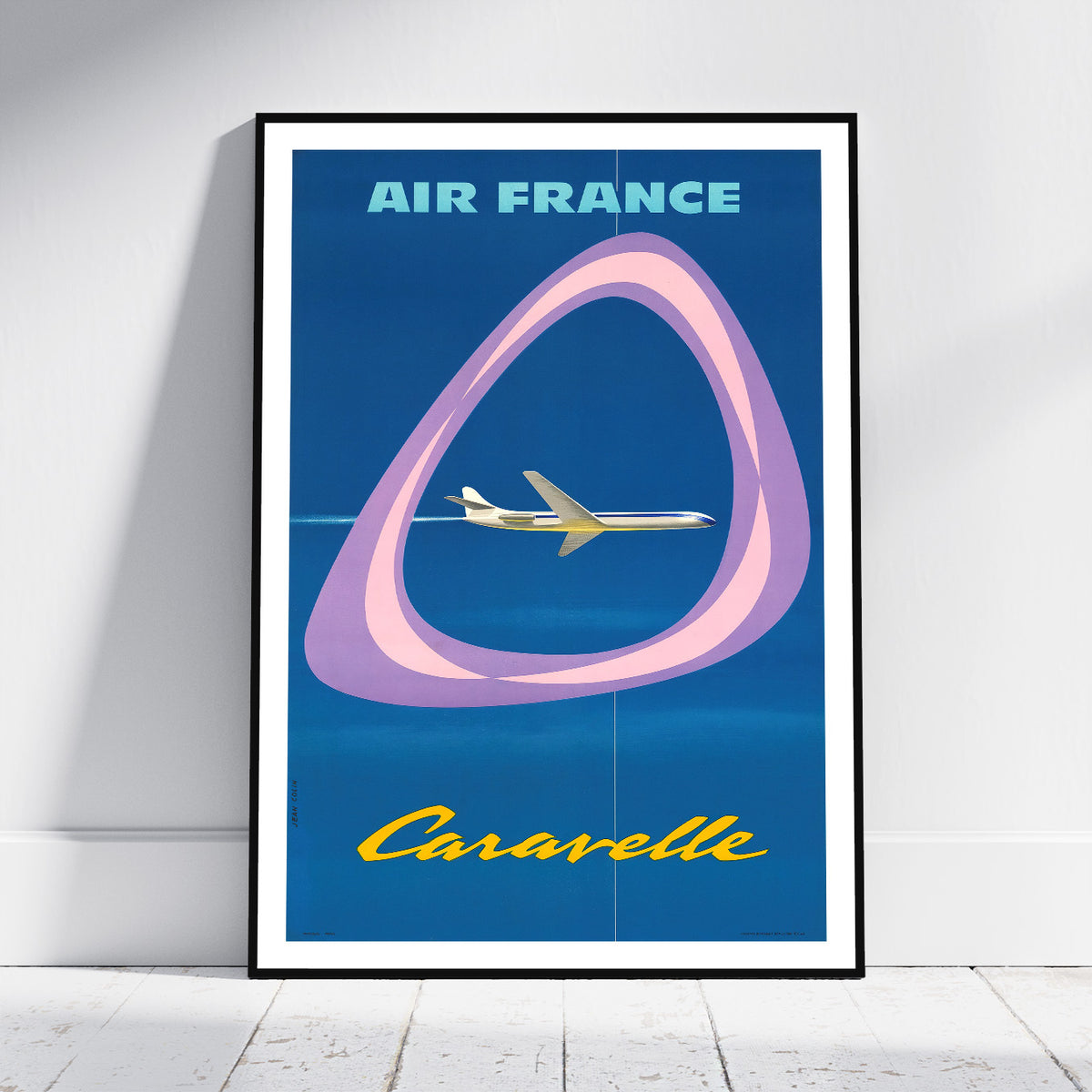 A framed vintage-style travel poster leaning against a white wall on a light floor. The poster reads 'AIR FRANCE' at the top and 'Caravelle' at the bottom, with a blue background, a pink and purple loop graphic, and a small airplane illustration.
