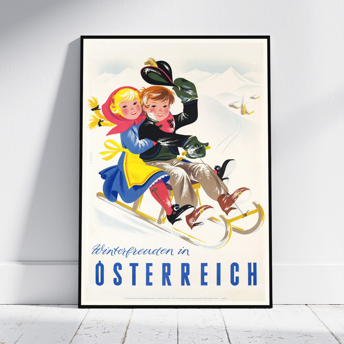 A framed vintage-style travel poster leaning against a white wall on a light wood floor. The poster shows two children sledding in a snowy mountain landscape with the text 'Winterfreuden in OSTERREICH' in blue at the bottom.