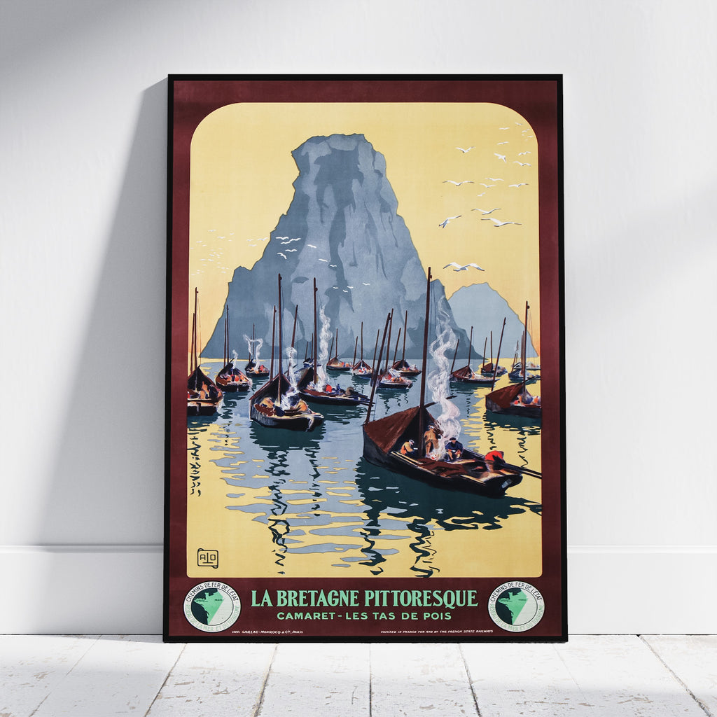 A framed vintage travel poster of La Bretagne Pittoresque, featuring sailboats on blue water in front of a large gray mountain under a pale yellow sky, leaning against a white wall on a light wood floor.