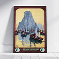 A framed vintage travel poster of La Bretagne Pittoresque, featuring sailboats on blue water in front of a large gray mountain under a pale yellow sky, leaning against a white wall on a light wood floor.