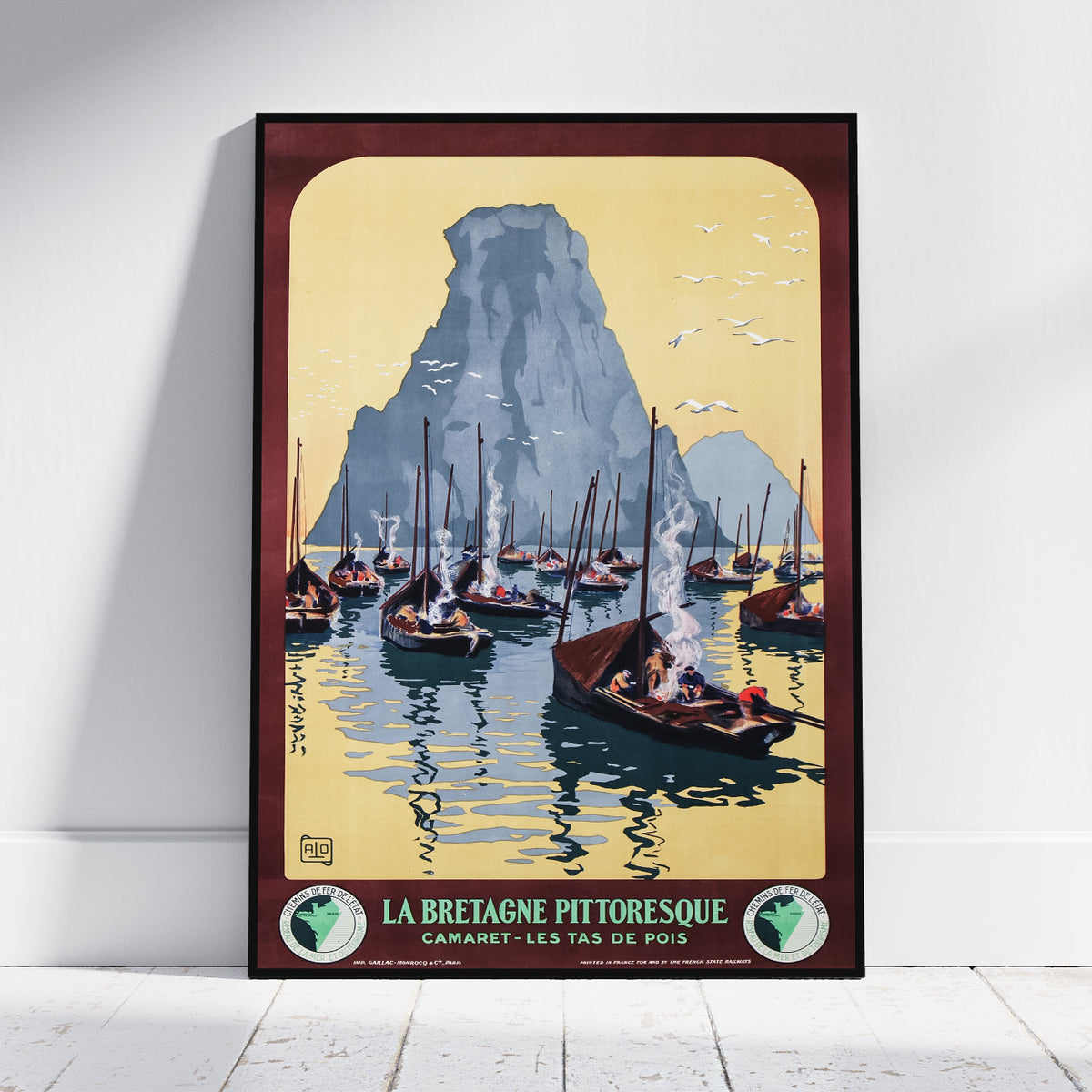 A framed vintage travel poster of La Bretagne Pittoresque, featuring sailboats on blue water in front of a large gray mountain under a pale yellow sky, leaning against a white wall on a light wood floor.