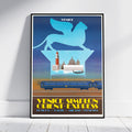 A framed vintage-style travel poster leaning against a white wall on a light wood floor. The poster features a blue winged lion silhouette above Venice landmarks, with a blue train on tracks and yellow text reading 'VENICE SIMPLON ORIENT-EXPRESS' and 'LONDRAN PARIS - MIANG - VENECIA'.