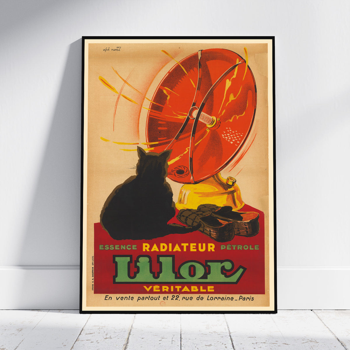 A framed vintage-style poster leaning against a white wall on a light floor. The poster shows a black cat in front of a large red radiator or fan-like object with yellow accents and bold text reading 'Essence Radiateur Petrole Ulor Veritable.