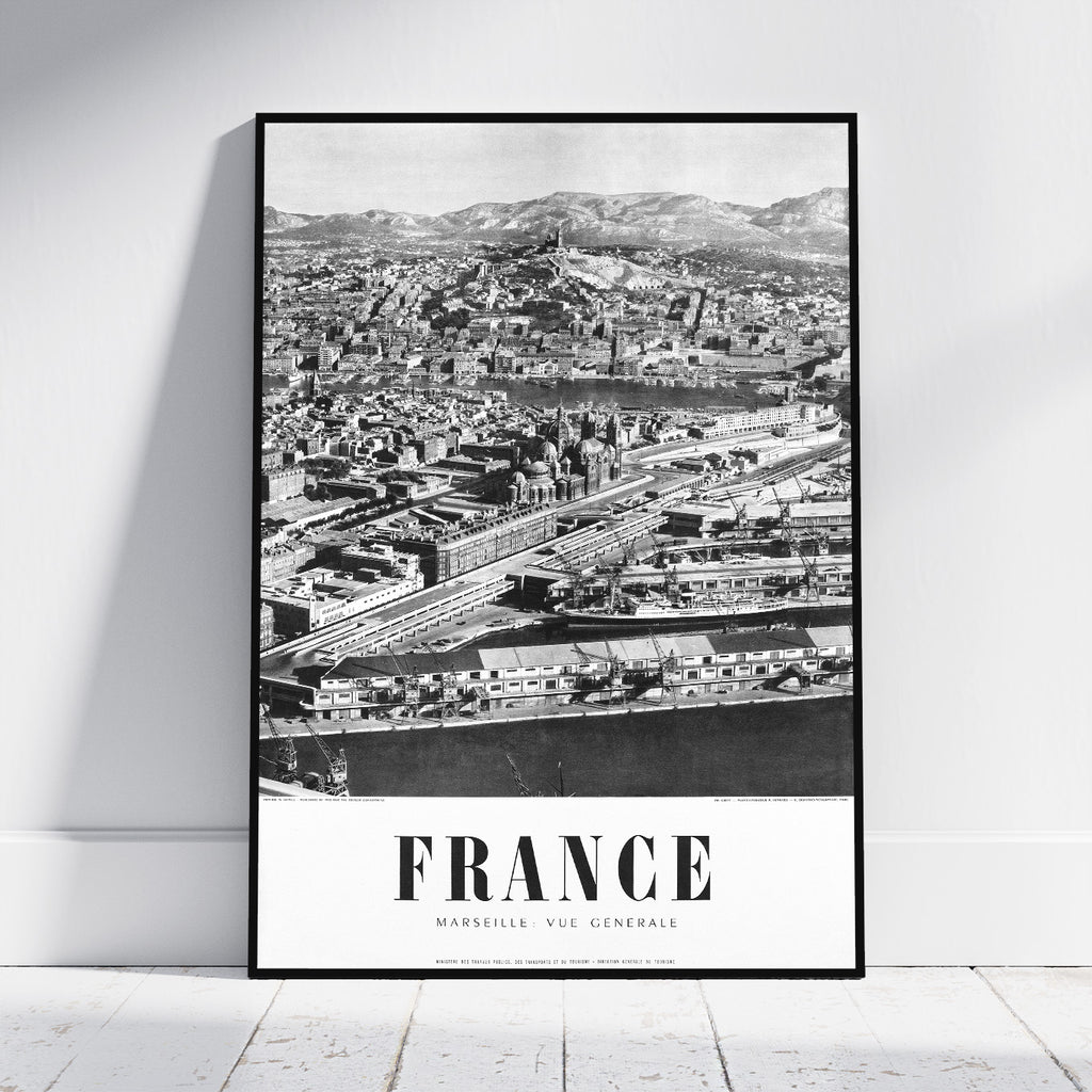 A framed black and white vintage-style poster of Marseille, France, leaning against a white wall on a light wooden floor.