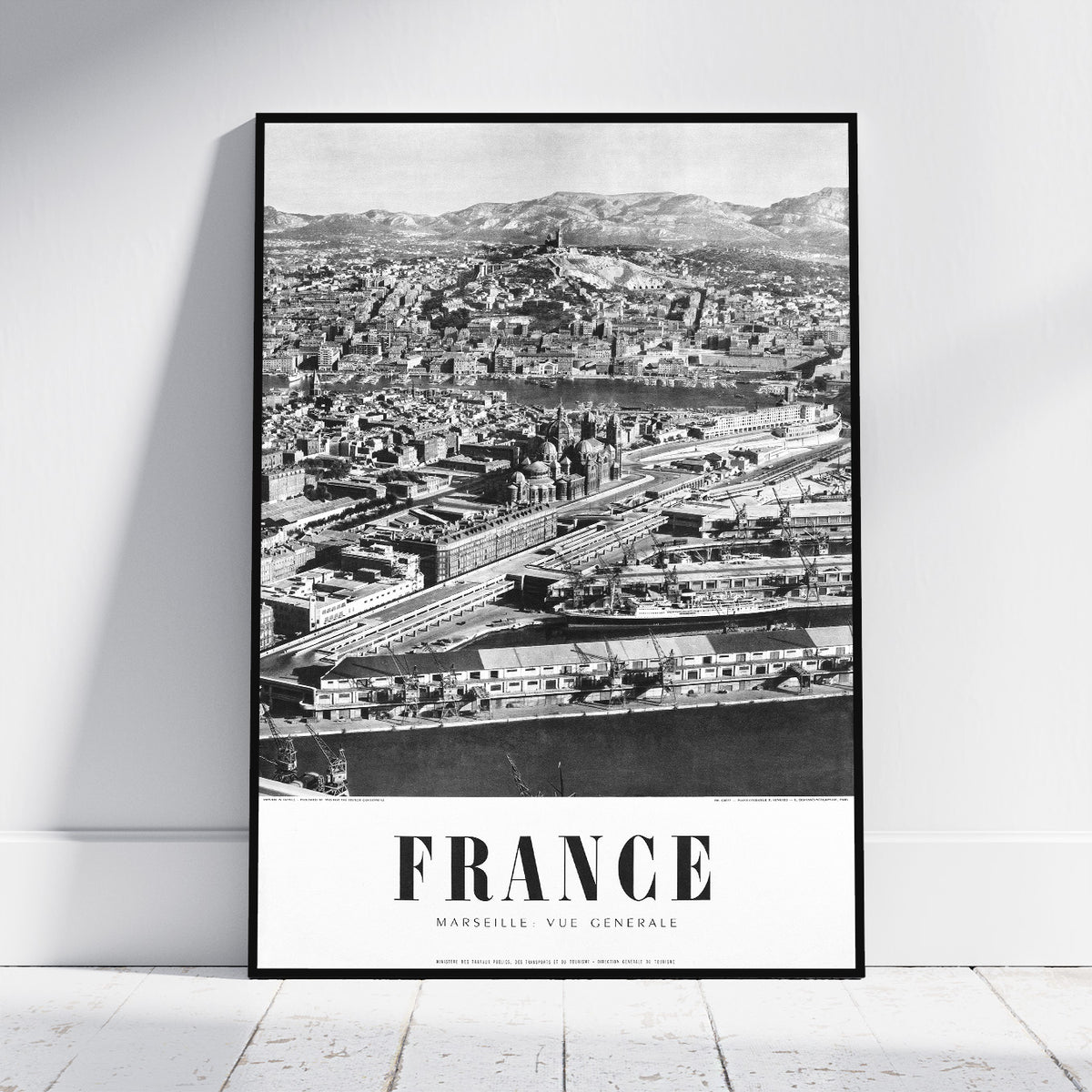 A framed black and white vintage-style poster of Marseille, France, leaning against a white wall on a light wooden floor.