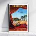 A framed vintage-style Air France travel poster leaning against a white wall on a light wood floor. The poster depicts a desert scene with palm trees, a seated figure under red drapery, an airplane in the sky, and the text 'AIR FRANCE' at the top and 'AFRIQUE DU NORD' at the bottom.