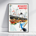 A framed vintage-style travel poster leaning against a white wall on a light wood floor. The poster features the text 'MESSAGERIES MARITIMES' in red at the top, an illustration of large cargo ships and smaller boats in a harbor, and a woman in a blue top in the lower right.