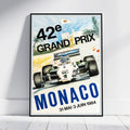 A framed vintage-style Monaco Grand Prix poster leaning against a white wall on a light floor. The poster shows a white Formula 1 race car on a track with large text reading '42èe GRAND PRIX', 'MONACO', and the dates '31 MAI - 3 JUIN 1984'.