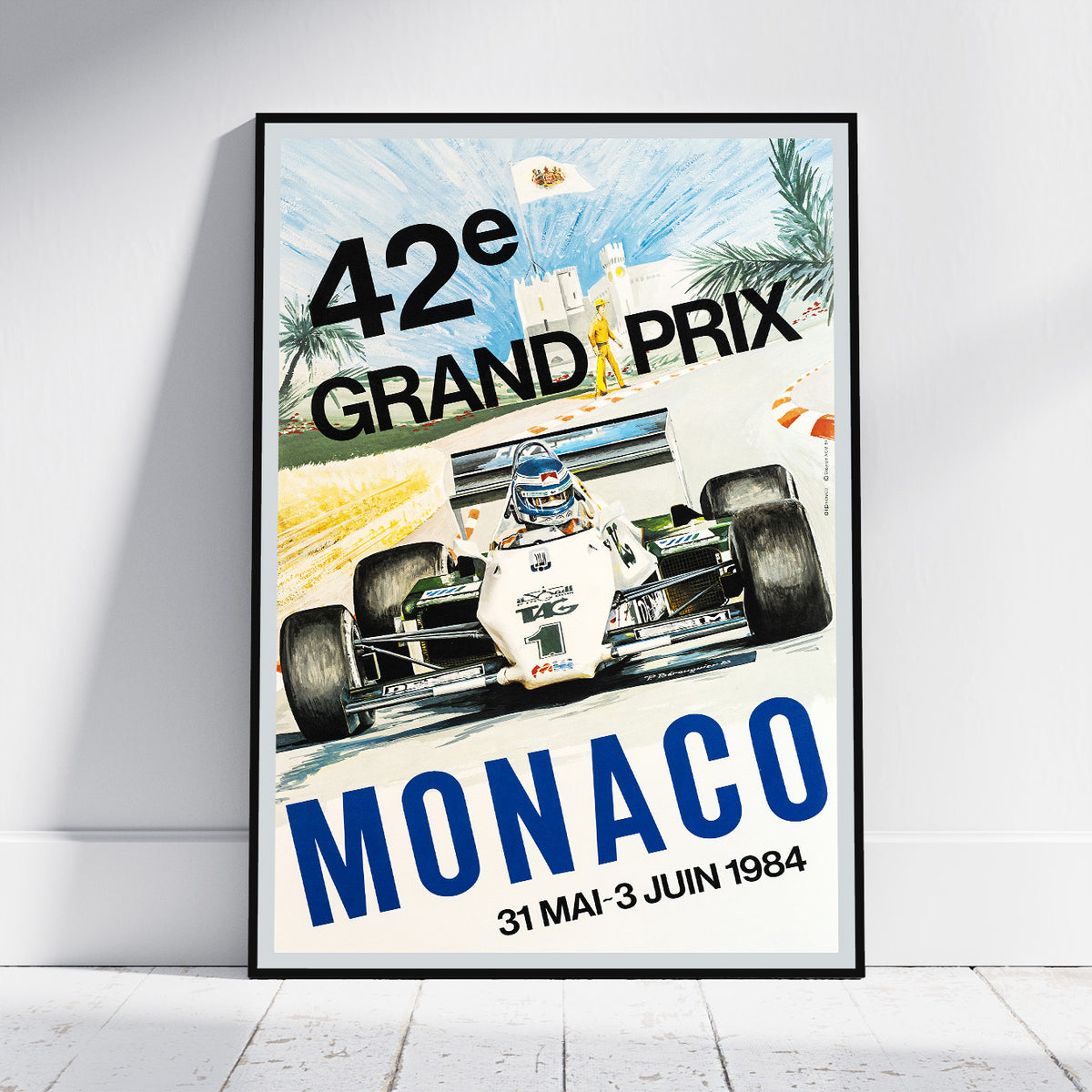 A framed vintage-style Monaco Grand Prix poster leaning against a white wall on a light floor. The poster shows a white Formula 1 race car on a track with large text reading '42èe GRAND PRIX', 'MONACO', and the dates '31 MAI - 3 JUIN 1984'.