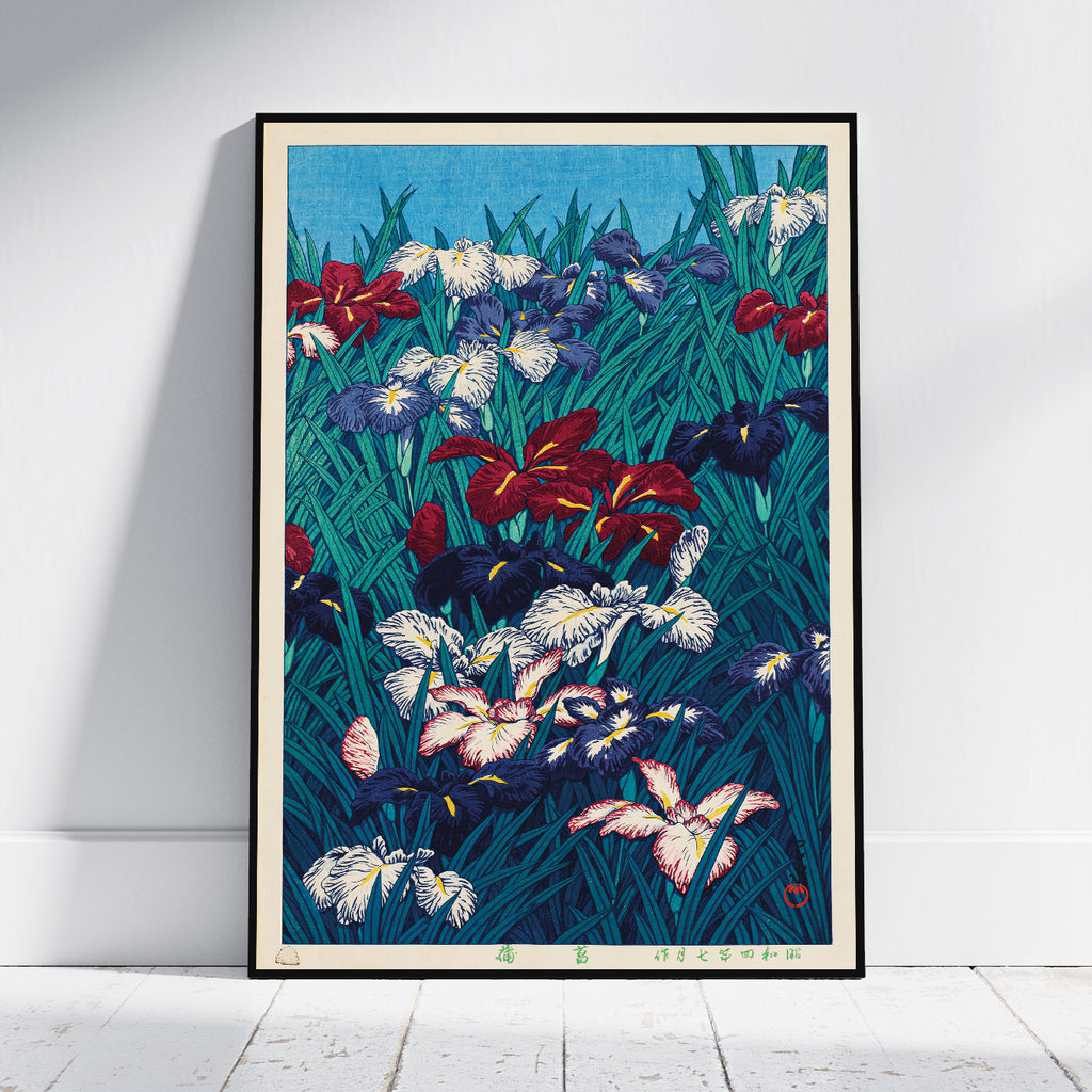 A framed art print of a Japanese-style iris garden, leaning against a white wall on a light wood floor. The print features a dense field of irises in red, white, purple, and blue on a dark teal background, enclosed in a thin black frame.