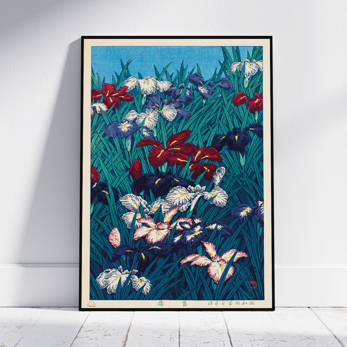 A framed art print of a Japanese-style iris garden, leaning against a white wall on a light wood floor. The print features a dense field of irises in red, white, purple, and blue on a dark teal background, enclosed in a thin black frame.