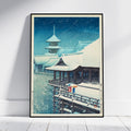 A framed art print depicting a traditional Japanese snow scene leaning against a white wall on a light wood floor.