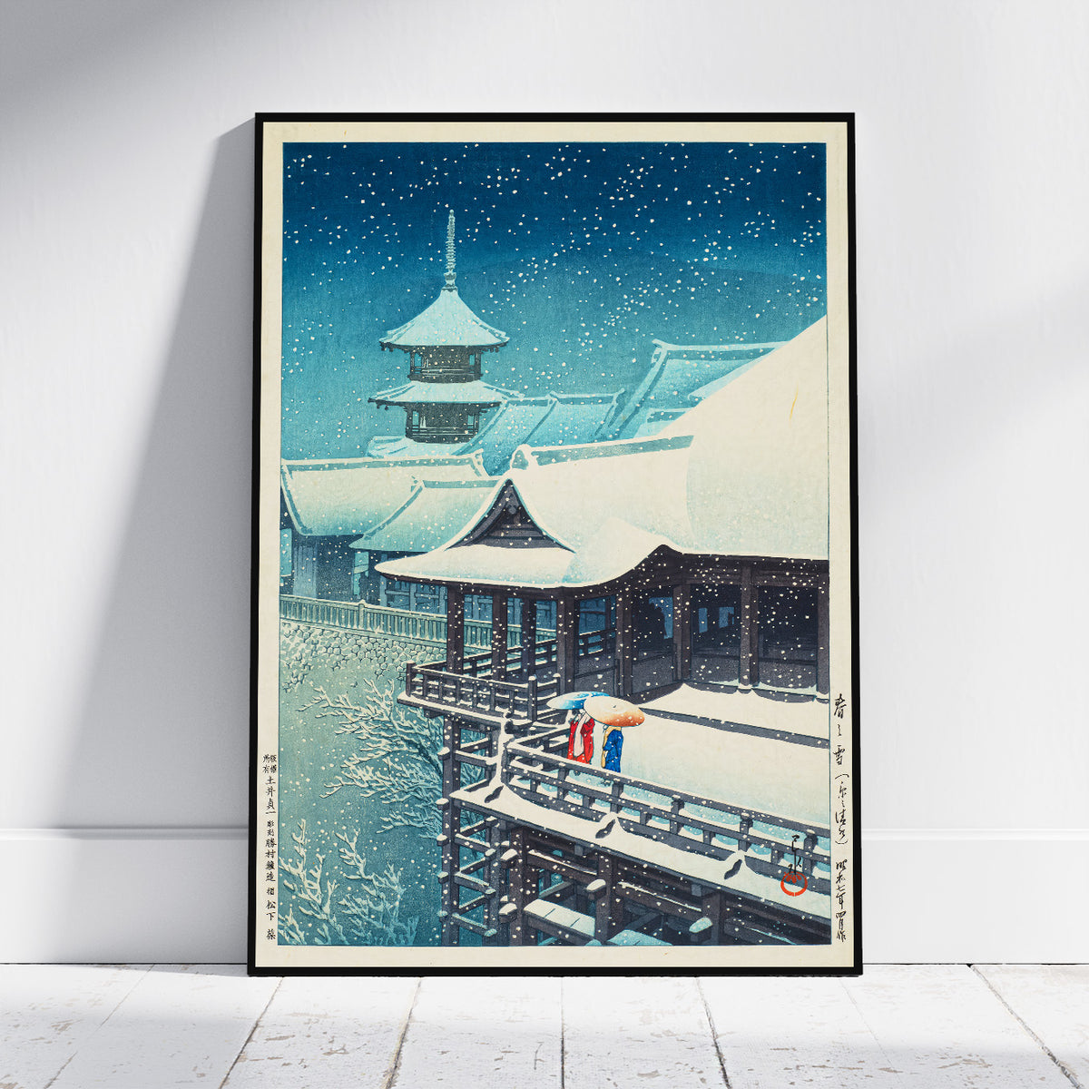 A framed art print depicting a traditional Japanese snow scene leaning against a white wall on a light wood floor.
