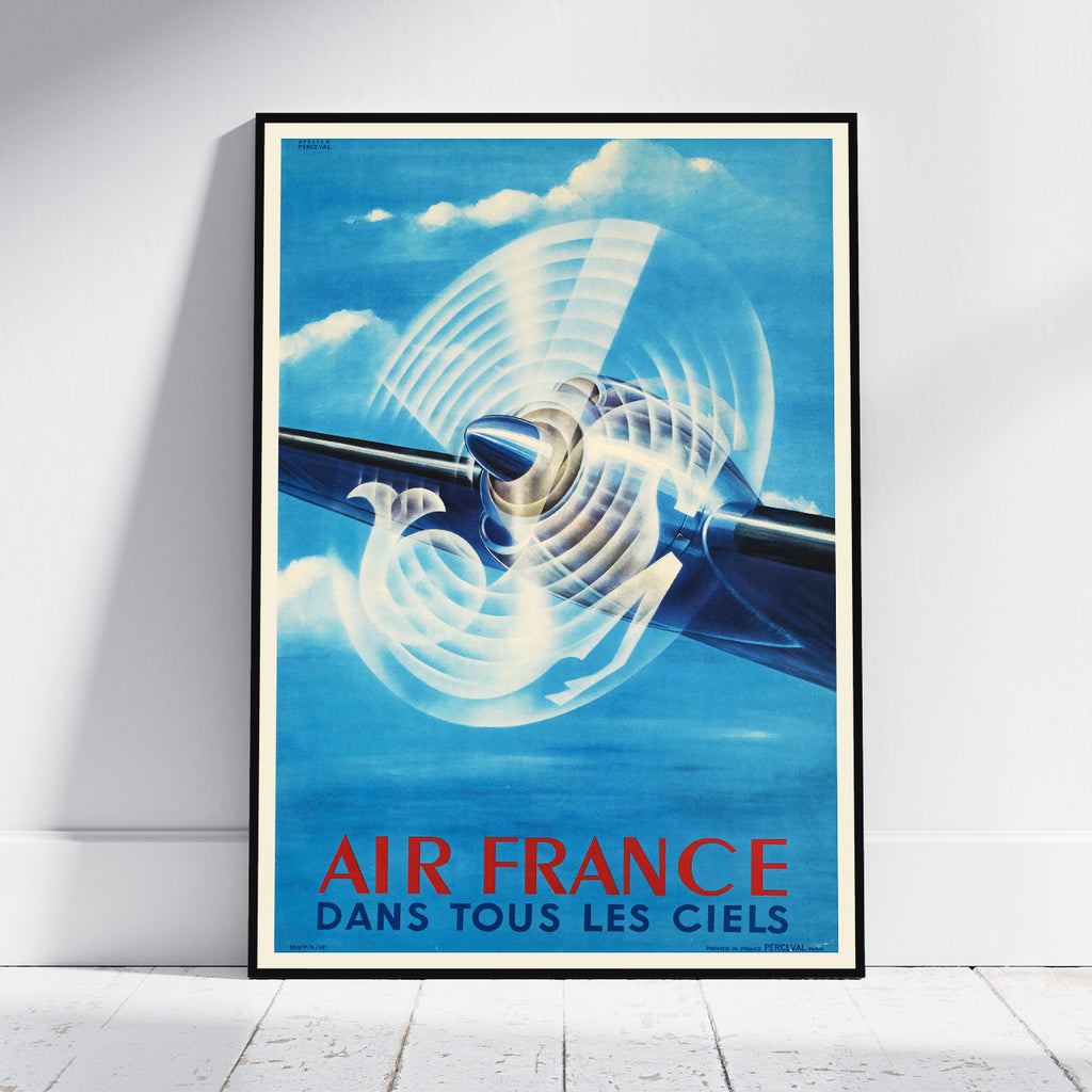 A framed vintage-style Air France poster leaning against a white wall on a light floor. The poster shows a blue airplane with a large white propeller and the text 'AIR FRANCE DANS TOUS LES CIELS' in red and blue at the bottom.