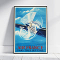 A framed vintage-style Air France poster leaning against a white wall on a light floor. The poster shows a blue airplane with a large white propeller and the text 'AIR FRANCE DANS TOUS LES CIELS' in red and blue at the bottom.