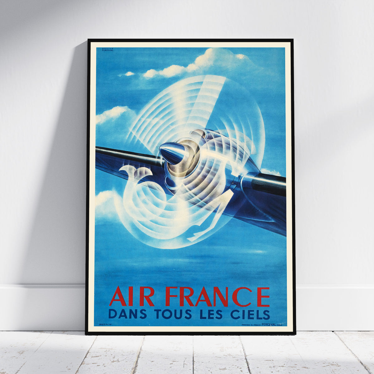 A framed vintage-style Air France poster leaning against a white wall on a light floor. The poster shows a blue airplane with a large white propeller and the text 'AIR FRANCE DANS TOUS LES CIELS' in red and blue at the bottom.