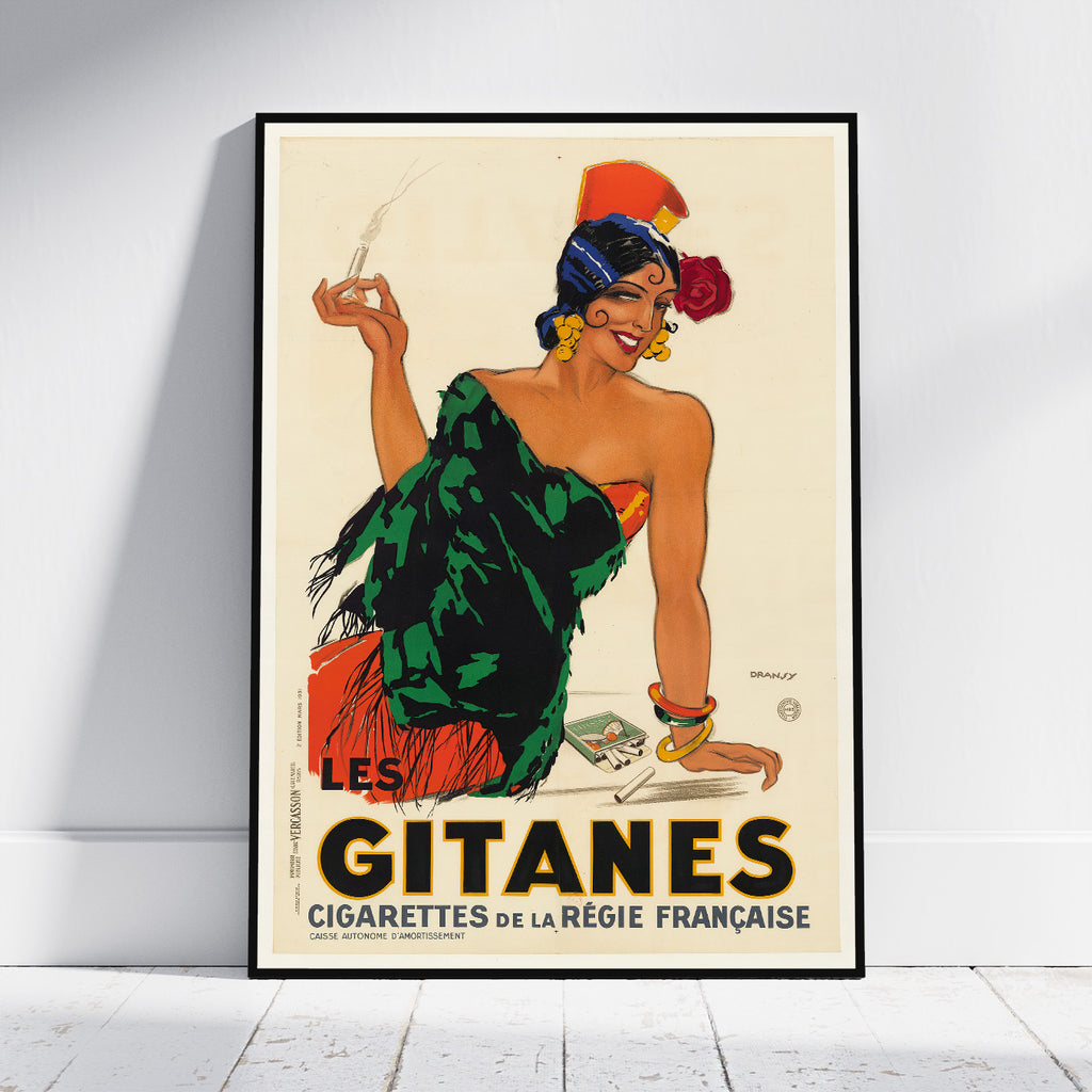 A framed vintage-style poster leaning against a white wall on a light floor. The poster shows an illustrated woman in a green and orange dress holding a cigarette, with large text reading “LES GITANES CIGARETTES DE LA RÉGIE FRANCAISE.