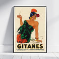 A framed vintage-style poster leaning against a white wall on a light floor. The poster shows an illustrated woman in a green and orange dress holding a cigarette, with large text reading “LES GITANES CIGARETTES DE LA RÉGIE FRANCAISE.
