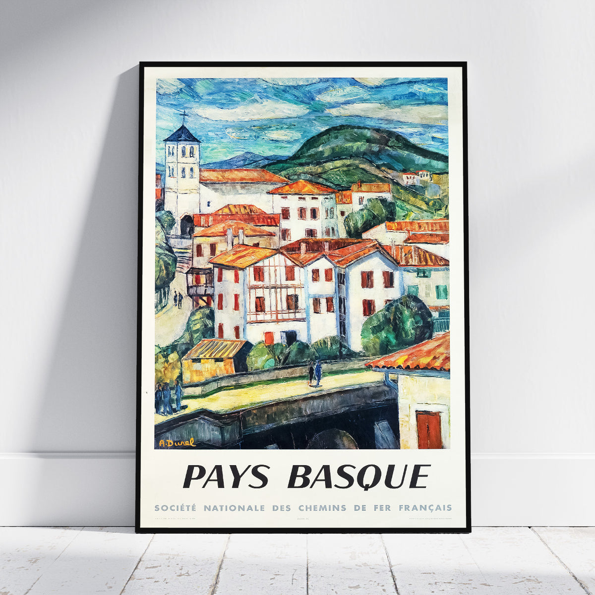 A framed travel poster print of a Basque village scene, leaning against a white wall on a light floor. The artwork shows colorful houses with orange roofs, green hills, and blue sky, with the text 'PAYS BASQUE' at the bottom.