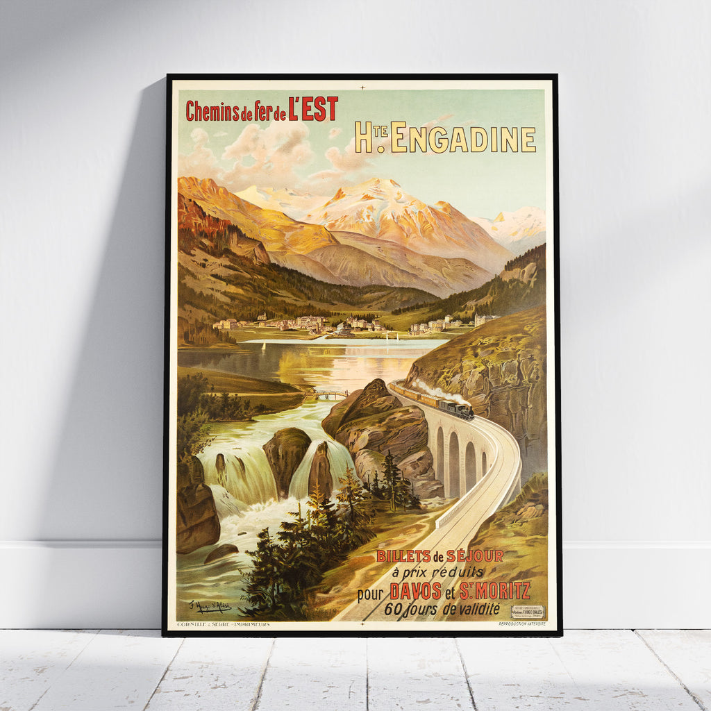 A framed vintage travel poster for Hè-Engadine, Switzerland, leaning against a white wall on a light wood floor. The poster features an illustrated mountain landscape with a train on a curved bridge over a river.