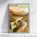 A framed vintage travel poster for Hè-Engadine, Switzerland, leaning against a white wall on a light wood floor. The poster features an illustrated mountain landscape with a train on a curved bridge over a river.