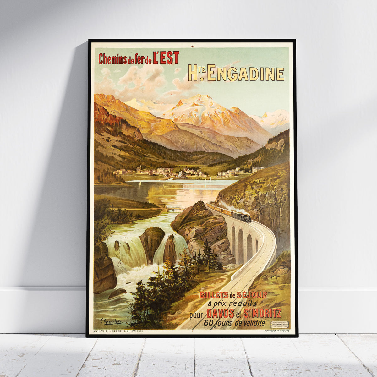 A framed vintage travel poster for Hè-Engadine, Switzerland, leaning against a white wall on a light wood floor. The poster features an illustrated mountain landscape with a train on a curved bridge over a river.