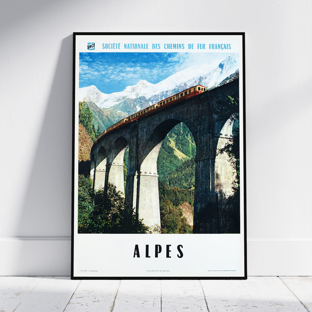 A framed vintage-style travel poster leaning against a white wall on a light wood floor. The poster shows a train traveling across a large stone viaduct in the Alps, with snow-capped mountains and green valleys under a blue sky.