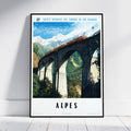 A framed vintage-style travel poster leaning against a white wall on a light wood floor. The poster shows a train traveling across a large stone viaduct in the Alps, with snow-capped mountains and green valleys under a blue sky.