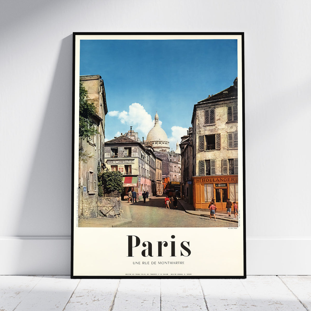 A framed travel poster of Paris, showcasing a vintage street scene with the Sacre-Coeur dome in the background under a blue sky. The poster features a cream-colored bottom border with the text 'Paris' and 'UNE RUE DE NOUVEAUTRICE'.