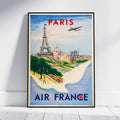 A framed vintage-style Paris travel poster leaning against a white wall on a light floor. The poster features the Eiffel Tower, Seine river, Paris skyline, an airplane, and large text reading 'PARIS' and 'AIR FRANCE'.