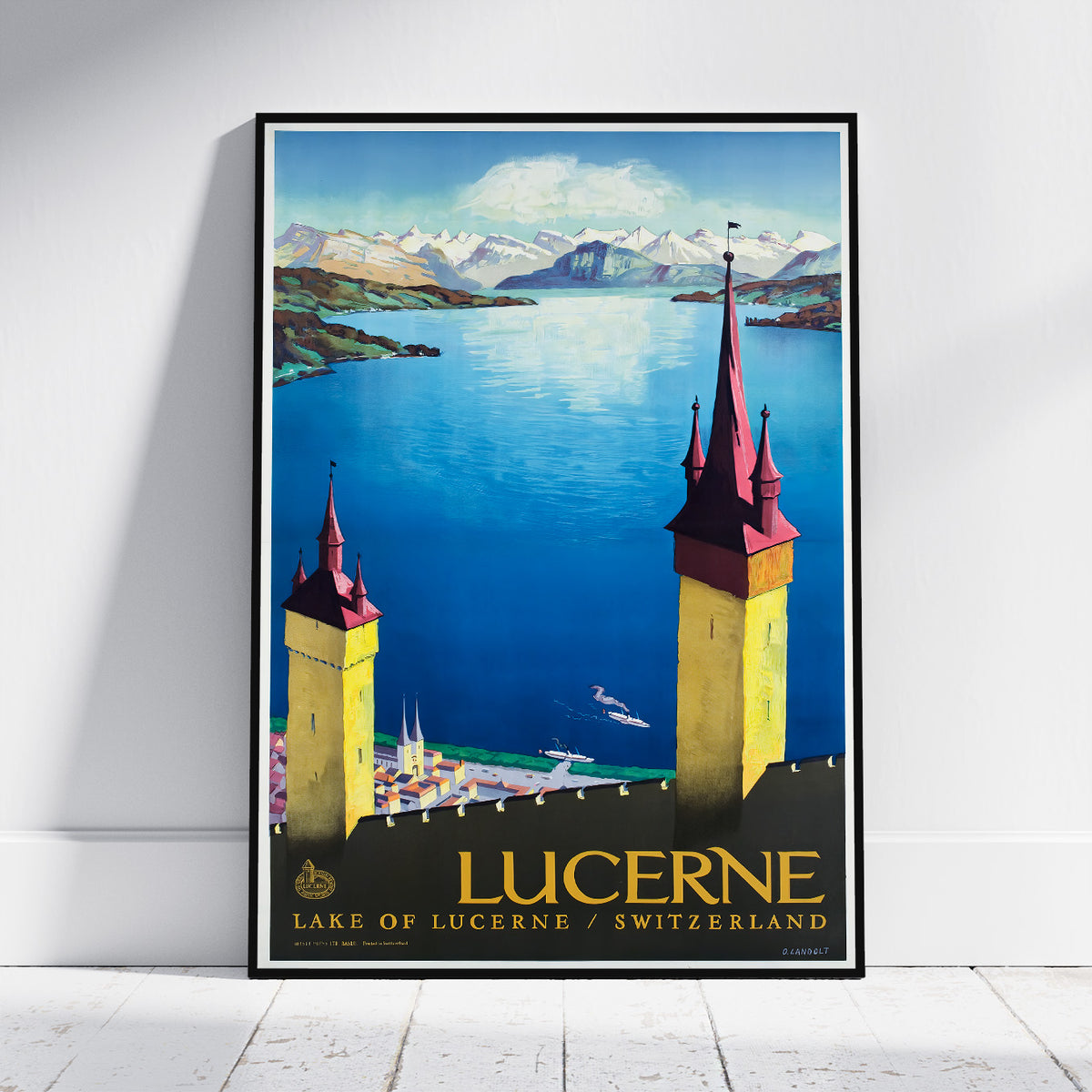 A framed vintage-style travel poster of Lucerne, Switzerland, leaning against a white wall on a light wood floor. The poster features a scenic illustration of the blue lake, snow-capped mountains, and two yellow towers with red roofs.