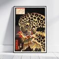 A framed Japanese-style art print leaning against a white wall on a light floor. The artwork shows a large spotted leopard or cat in profile with a red bird in its mouth, rendered in black, gold, cream, and red tones with Japanese calligraphy in the upper left and a red vertical label near the lower left.