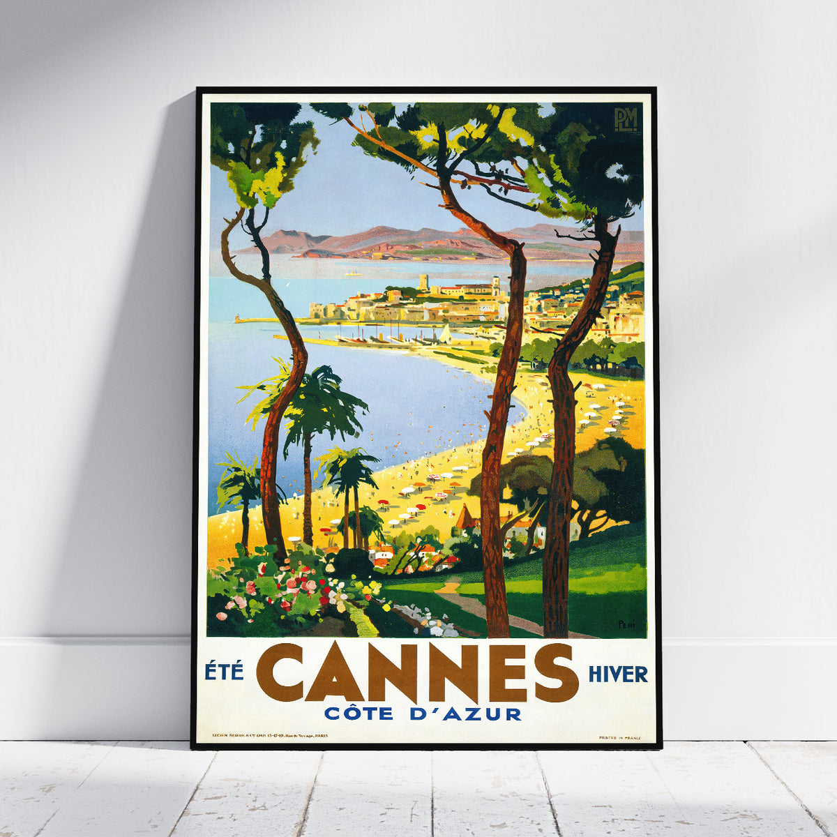 A framed vintage-style travel poster of Cannes, France, leaning against a white wall on a light wood floor. The poster features a colorful coastal landscape with trees, a bay, beach, and the text 'Cannes Cote d'Azur'.