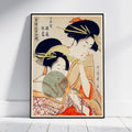 A framed Japanese ukiyo-e woodblock print depicting two women in traditional attire, leaning against a white wall on a light wood floor.